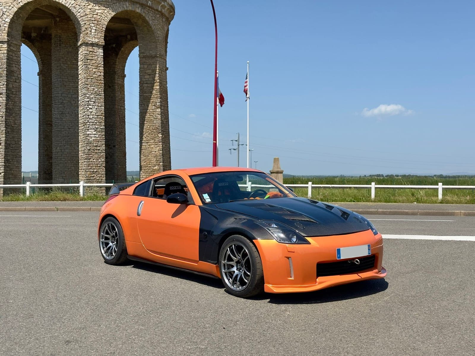 Nissan 350Z orange avec capot carbone protégé par film XPEL, vue trois-quarts avant devant la tour d'eau près du circuit de Magny-Cours